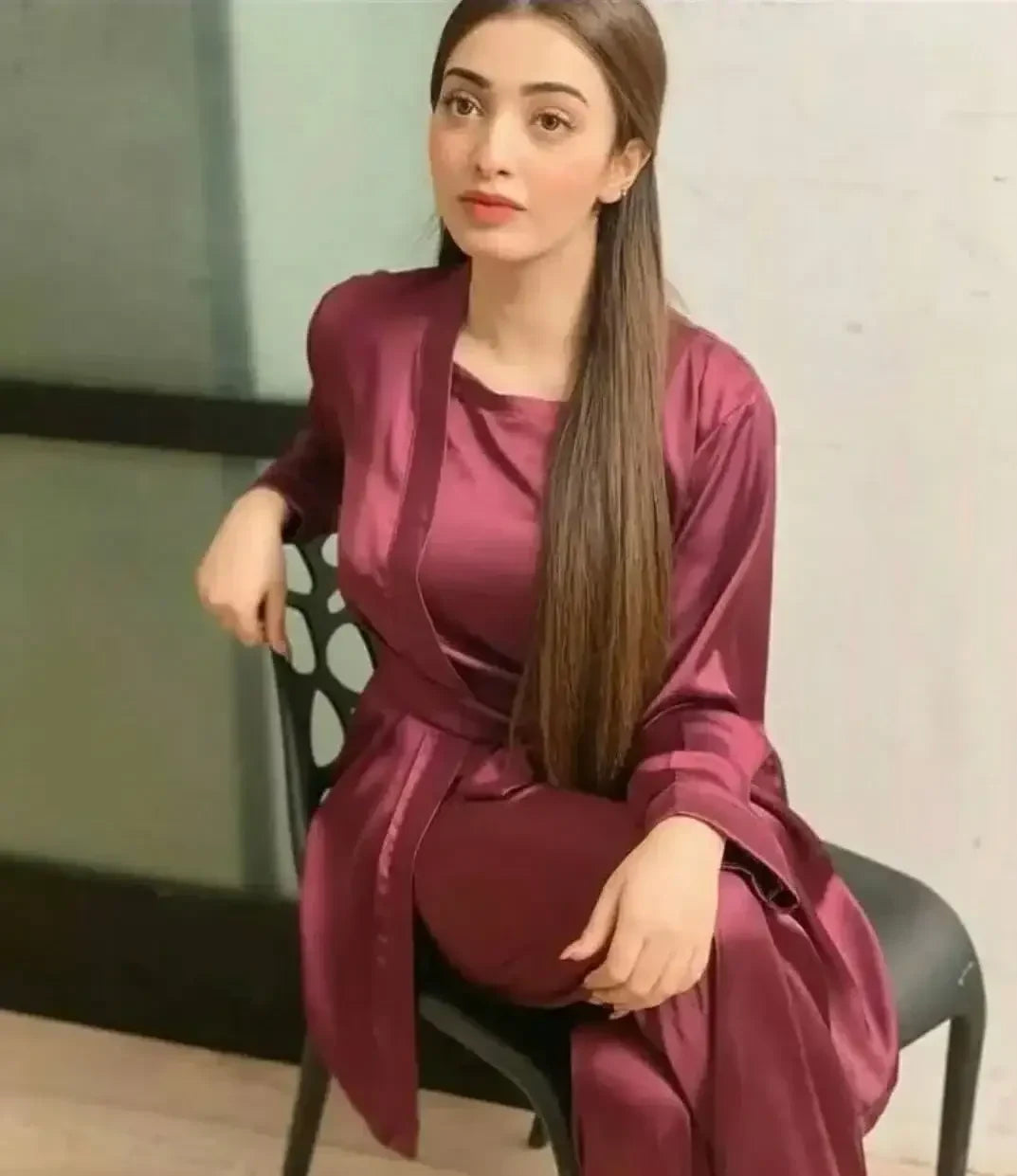 Young woman with long straight hair sitting on black chair wearing shiny maroon satin outfit against neutral background