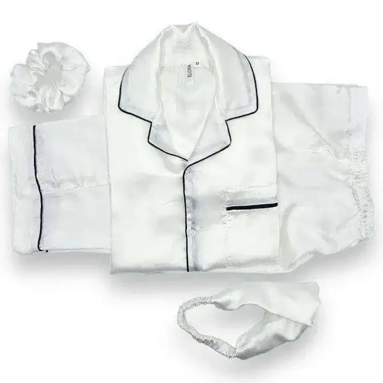 White silk pajamas with black piping, accompanied by a matching silk scrunchie and eye mask
