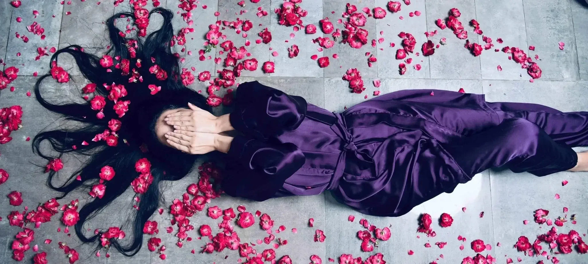Woman lying on tiled floor in purple silk robe with long black hair spread with pink flower petals